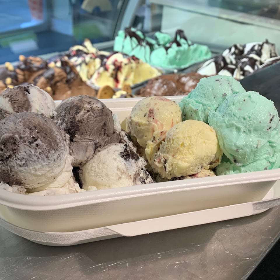 A white container filled with scoops of mint, vanilla, and chocolate gelato, with more in the background.