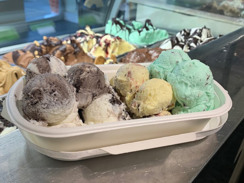 A white container filled with scoops of mint, vanilla, and chocolate gelato, with more in the background.
