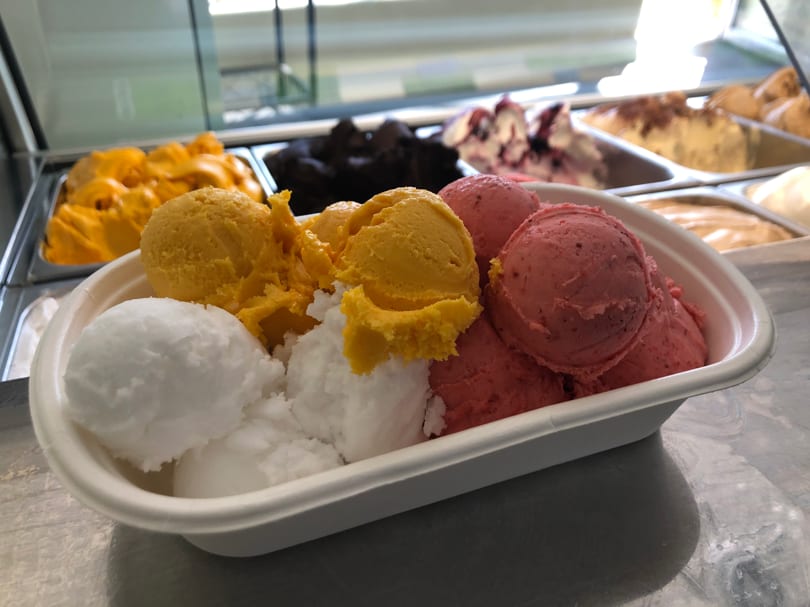 A white container filled with scoops of white, yellow, and pink gelato, with more flavors in the background.